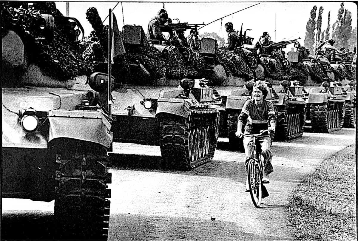 1985-11-Zoom-Dino-Fracchia-foto-bici-guerra-02 Carovane di carri armati durante un corteo storico, donna in bicicletta in primo piano, scena di protest and memoria storica.
